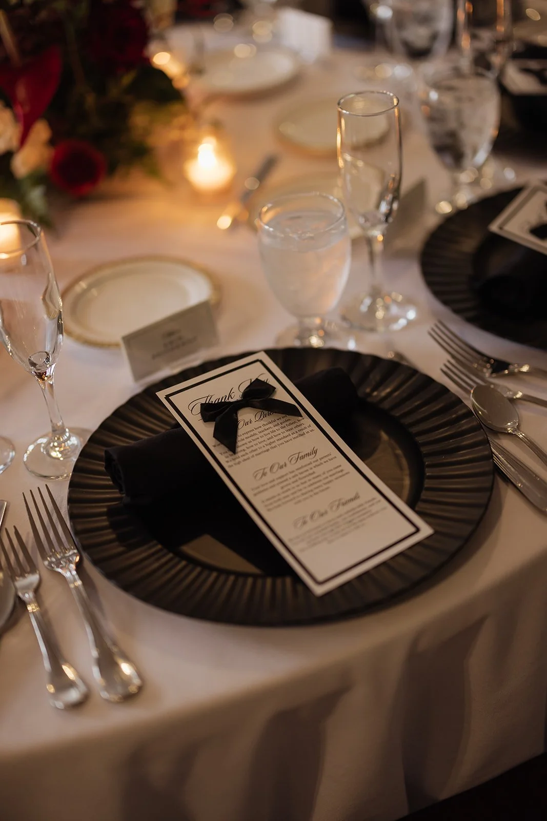 Elegant banquet table setting with black and white theme, featuring a black charger plate, a black napkin with a ribbon, a menu card, and multiple wine and water glasses, with warm lighting and floral centerpiece in the background.
