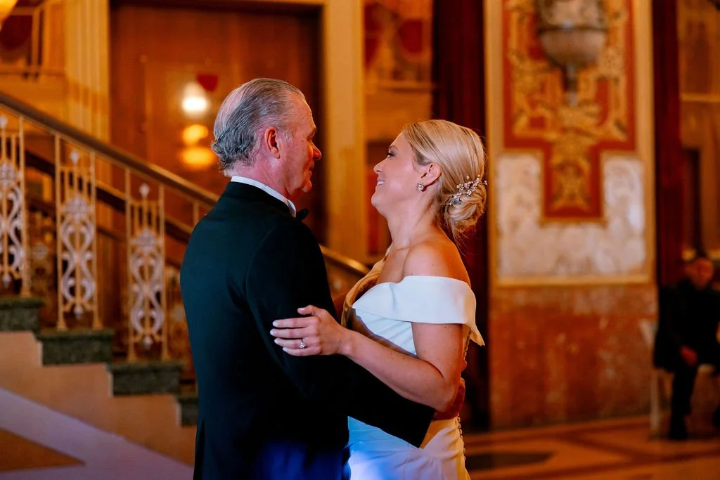 A bride and an older man are dancing and smiling at a wedding reception in a grand, warmly lit hall with wooden decor and ornate artwork.