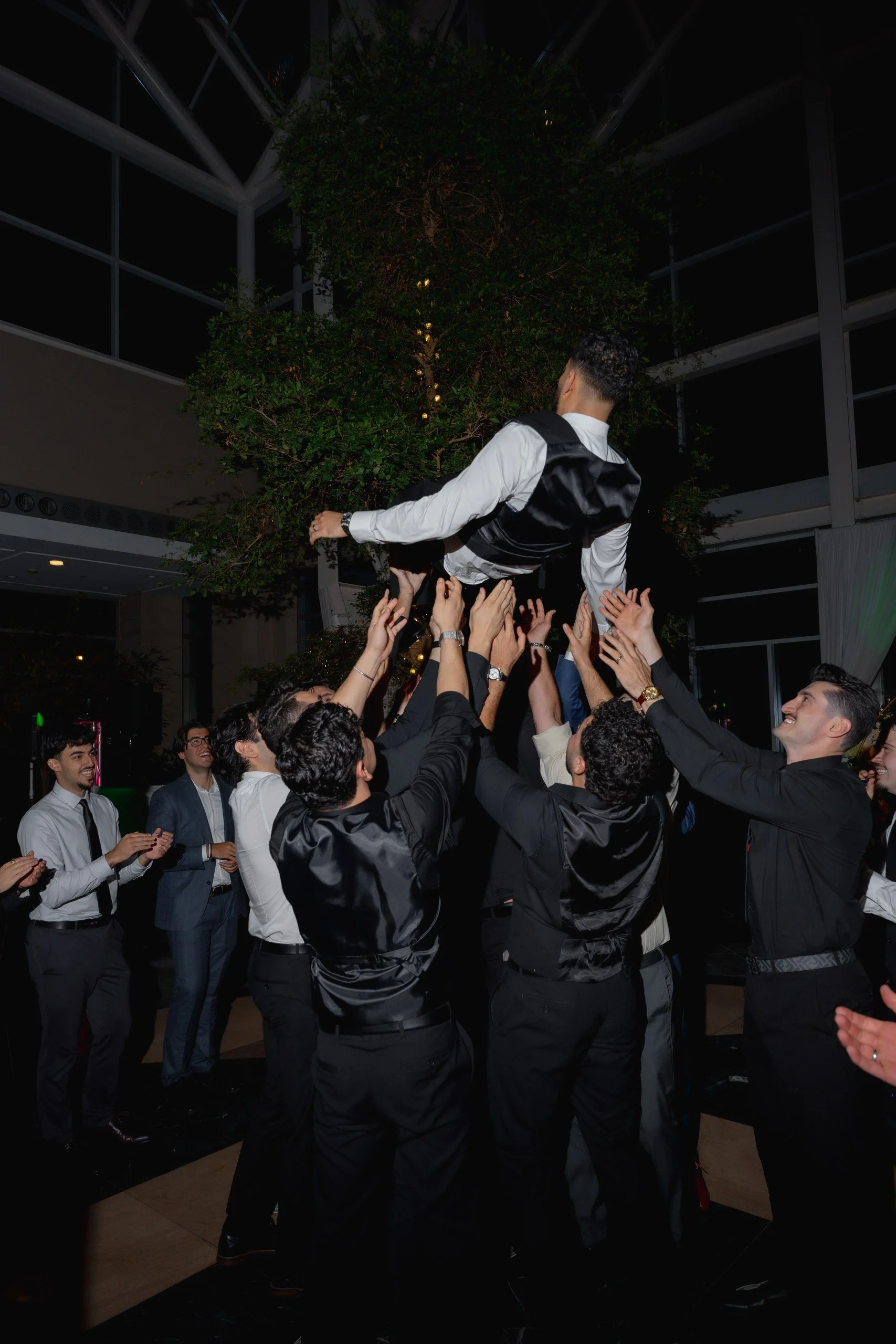 A group of men, dressed in formal attire, are celebrating at a party by lifting a man in a white shirt and black vest into the air. The setting appears to be indoors with a modern architectural design and a large indoor tree in the background.