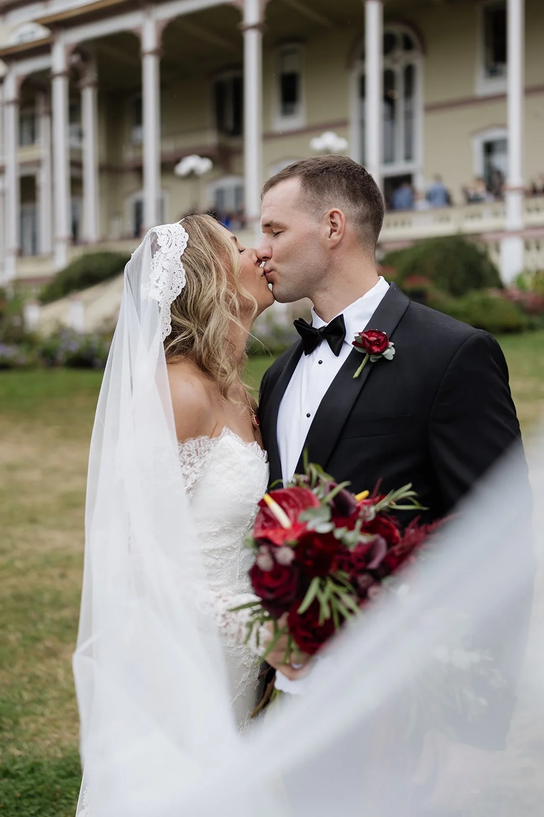 A bride and groom share a kiss outdoors on their wedding day, with a large house in the background. The bride is wearing a white wedding dress and a veil, and the groom is dressed in a black tuxedo with a red boutonniere. The bride holds a bouquet of