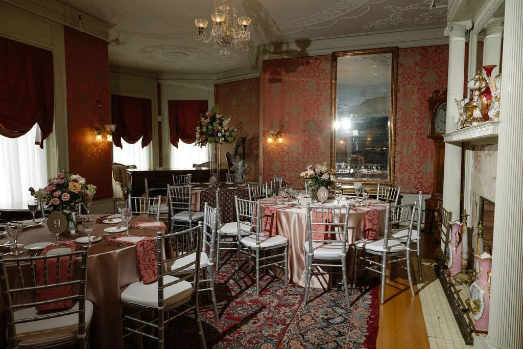 Elegant dining room with round tables covered in pink satin tablecloths, decorated with floral centerpieces, set with glassware and pink napkins, surrounded by silver chairs. The room features ornate wallpaper, a large mirror, a chandelier, and a fir