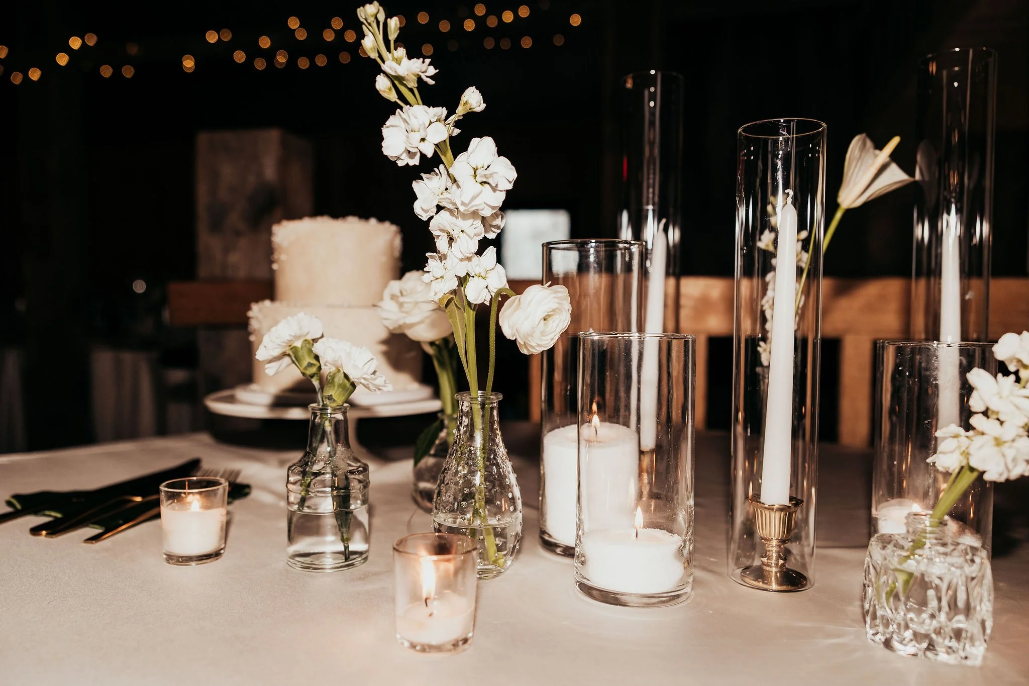 Elegant table decorated with white flowers in glass vases, white candles in holders, and tall glass candleholders, with a blurred background of string lights.