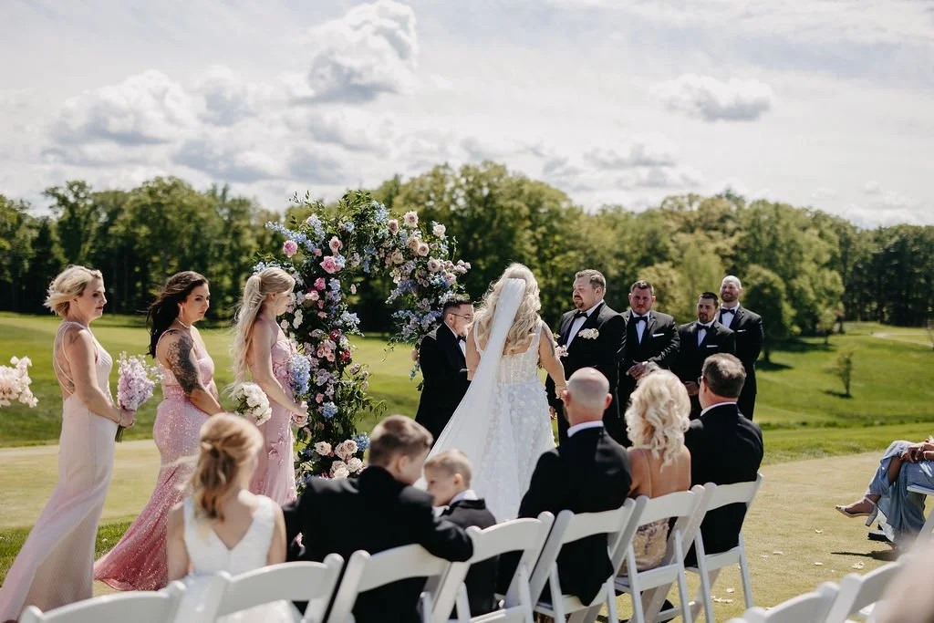 A wedding ceremony outdoors on a sunny day with the bride and groom standing under a floral arch, surrounded by bridesmaids, groomsmen, and guests seated on white chairs on a lush green lawn with trees and a cloudy sky in the background.
