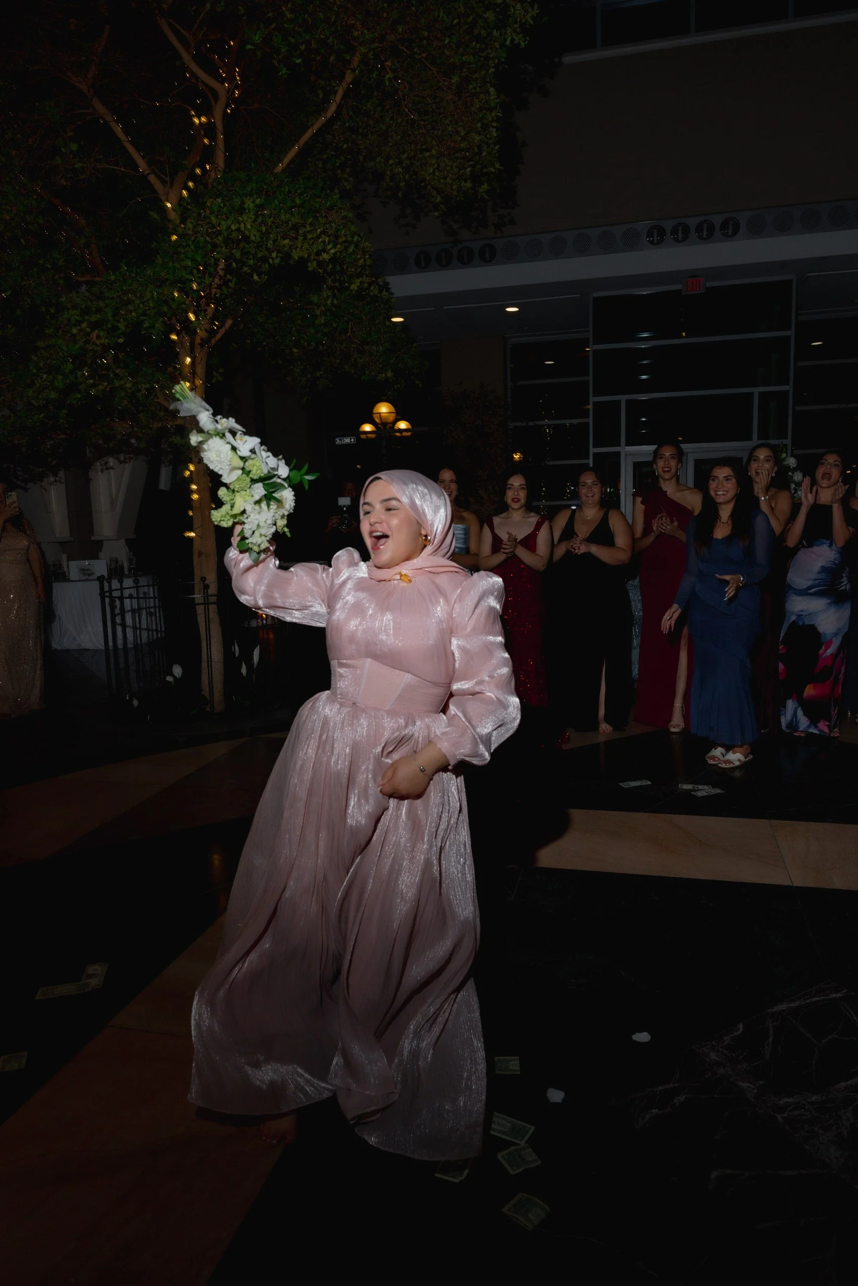 A woman in a pink gown holding a bouquet of white flowers, celebrating at an indoor event with other women in elegant dresses in the background, some with hands clapping or covering mouths, at night with outdoor lighting and trees.