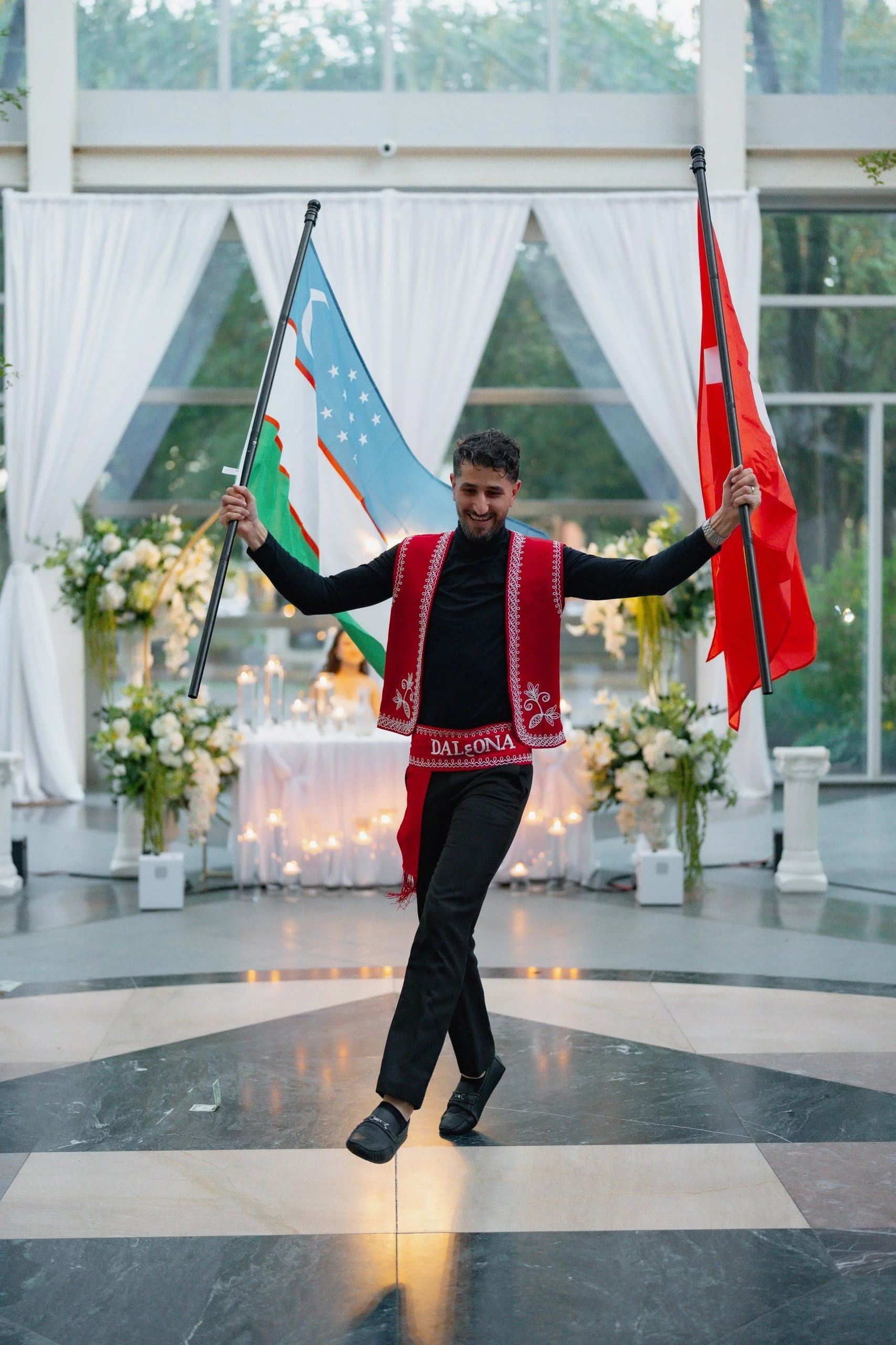 A man dancing indoors holding two flags, one blue and one red, in a decorated room with white curtains, floral arrangements, and candlelit tables.