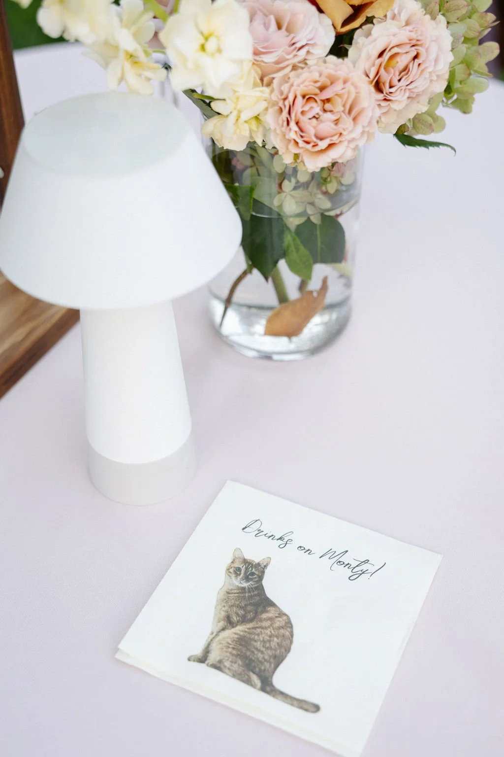 A white lamp, a glass vase with pink and cream flowers, and a greeting card with a cat illustration and the handwritten message 'Thanks on Monday!' on a light-colored surface.