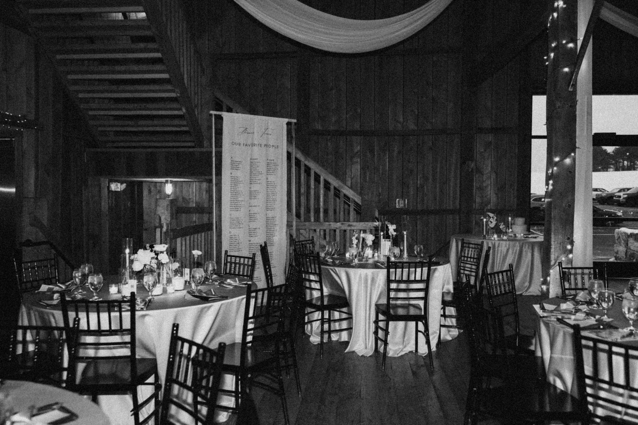 Indoor event space with round tables covered in white tablecloths, decorated with flowers and glassware, arranged for a formal occasion. Dark wood-paneled walls, a staircase, and string lights are visible, with a large window showing parked cars outs