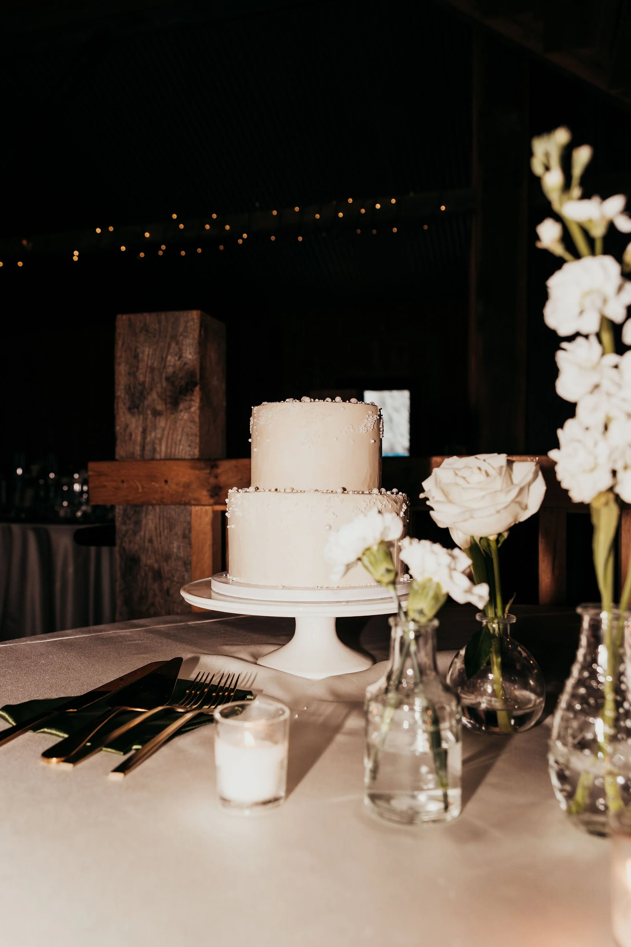 White tiered wedding cake with pearl decorations on a white cake stand, surrounded by white flowers in glass vases and candles on a table, with a rustic wooden wall background and soft lighting.