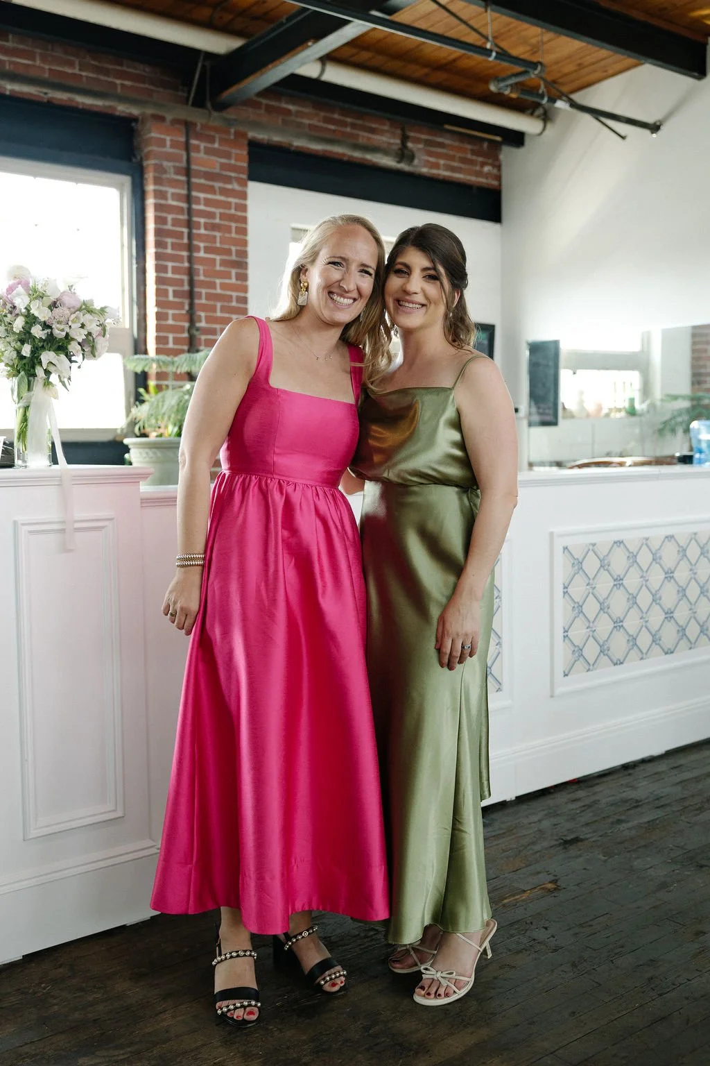 Two women in formal dresses smiling inside a decorated venue, one in a bright pink gown and the other in an olive green dress, standing close together with arms around each other.