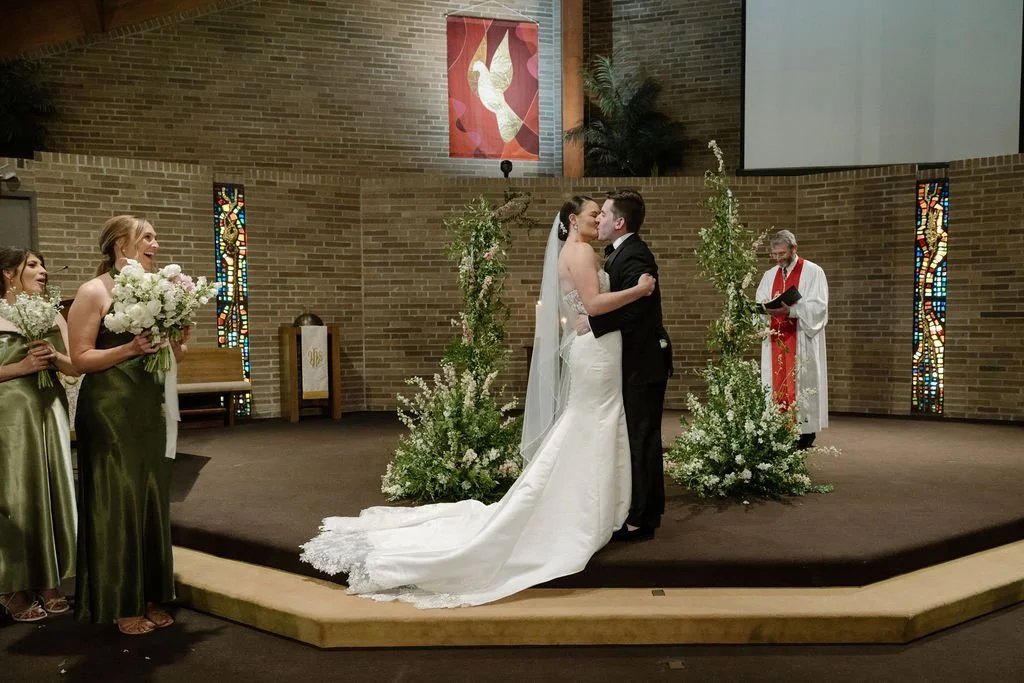 A newlywed couple sharing their first kiss in a church during their wedding ceremony. The bride is in a white gown with a long train and veil, and the groom is in a black tuxedo. Bridesmaids in green dresses holding bouquets stand to the left, while 
