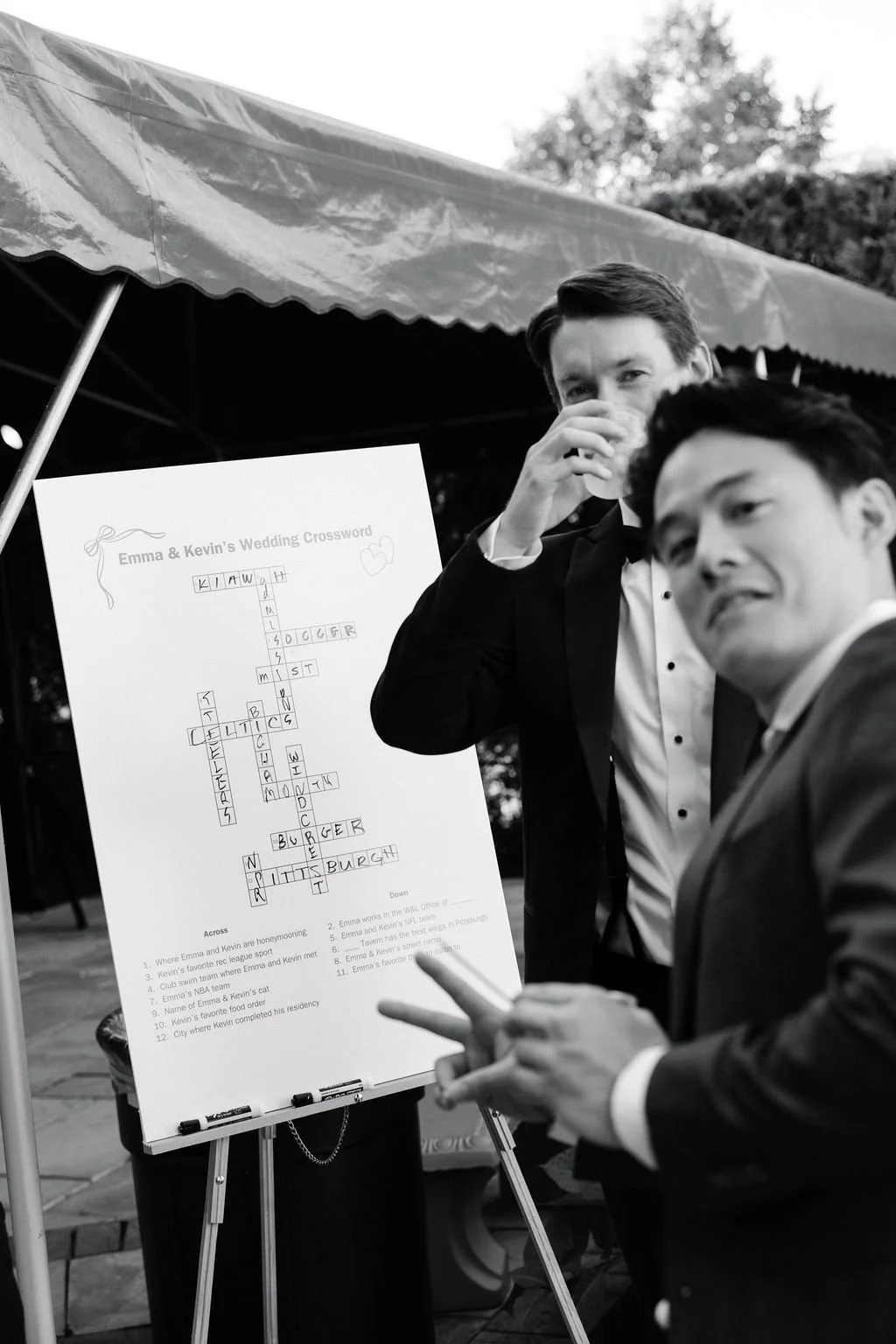 Two men in tuxedos at a wedding reception looking at a crossword puzzle on a whiteboard under a canopy. The crossword is titled 'Emma & Kevin's Wedding Crossword.' One man is holding up two fingers, and the other is covering his nose.