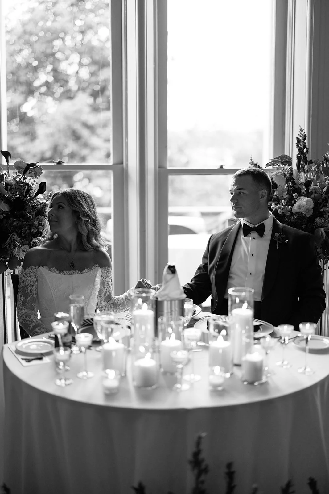 A couple at a wedding reception, sitting at a table with lit candles, in front of large windows with outdoor scenery visible.