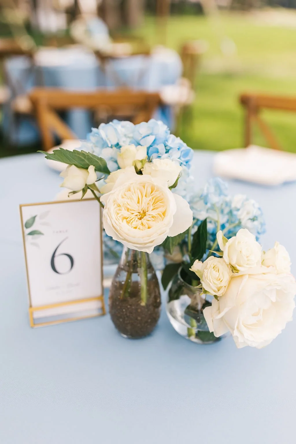 Floral centerpiece with white and light blue flowers on a table at an outdoor event, with a table number card displaying the number 6.