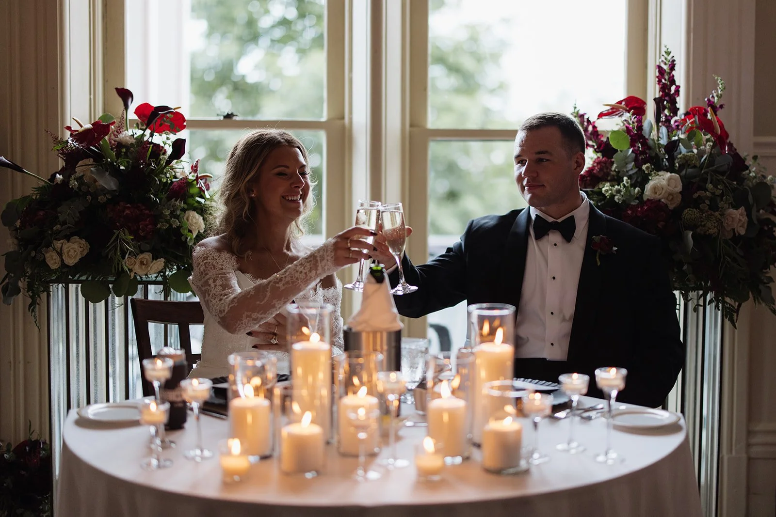 Bride and groom celebrating with champagne at a wedding reception, seated at a table with lit candles and floral arrangements in the background.