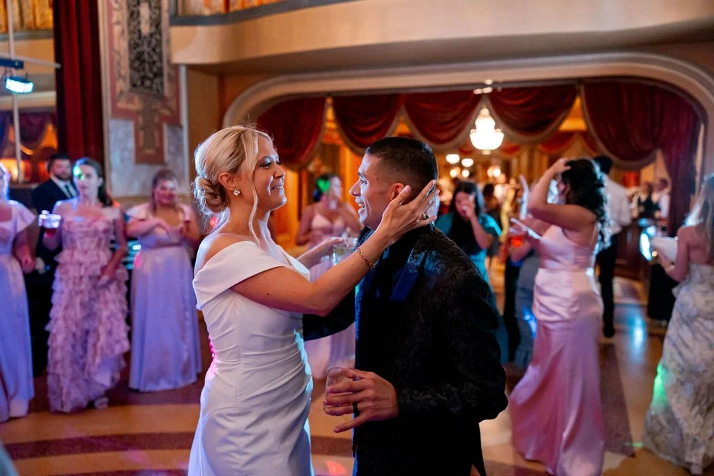 A bride and groom dancing at their wedding reception, surrounded by guests in formal attire, inside a decorated banquet hall.
