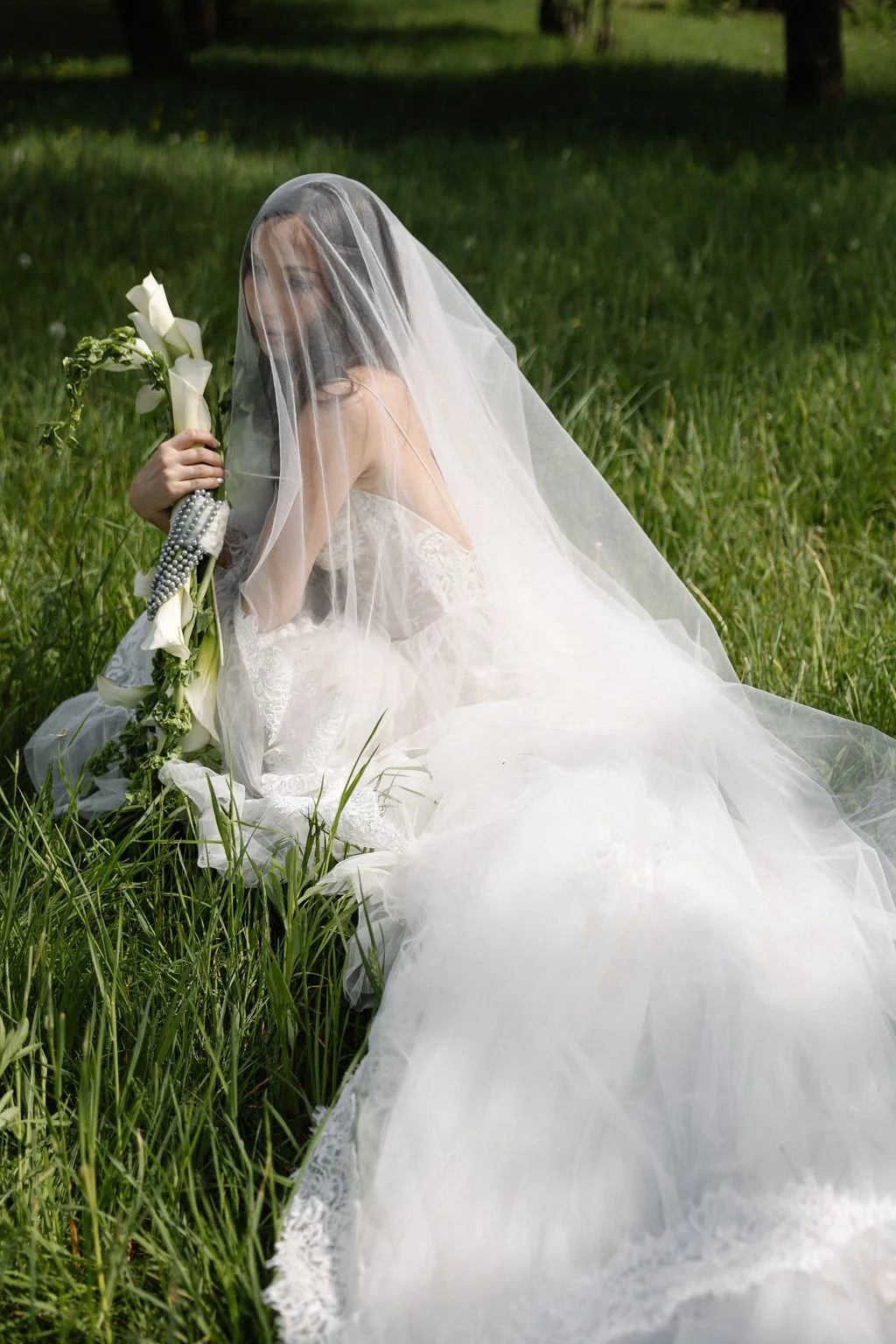 A bride in a white wedding gown sitting on grass in a green field, holding a bouquet of white calla lilies, with her face partially covered by a veil, surrounded by tall grass and trees in the background.