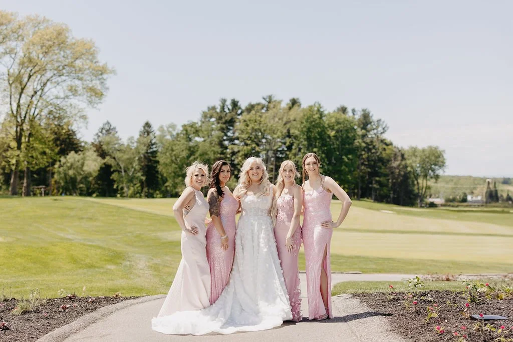 Five women in formal gowns standing together on a golf course with green trees and blue sky in the background