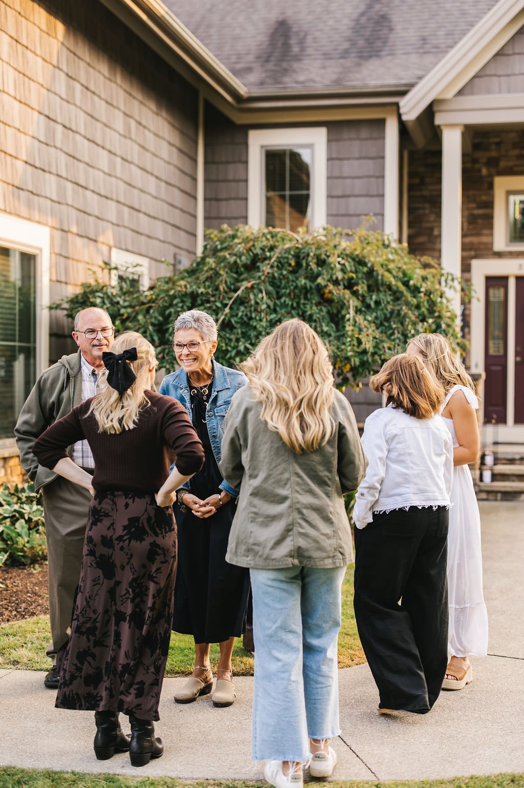 Group of six people talking and smiling outside a house with tan siding and steps.
