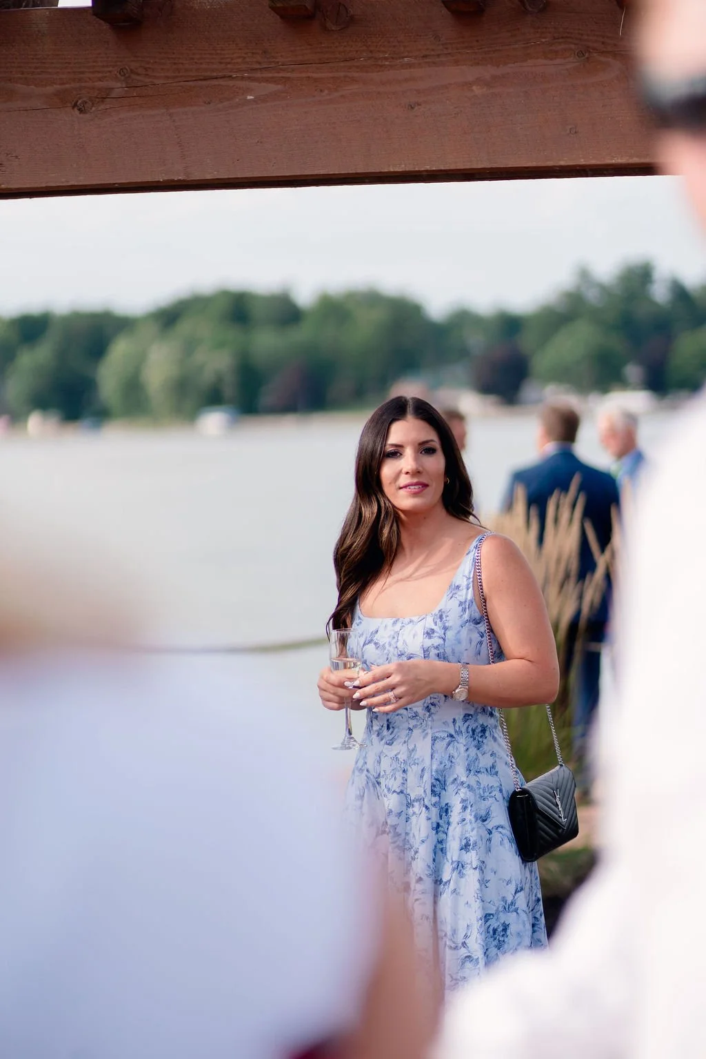 A woman in a light blue floral dress holding a wine glass at an outdoor social event near a body of water, with other people in the background.