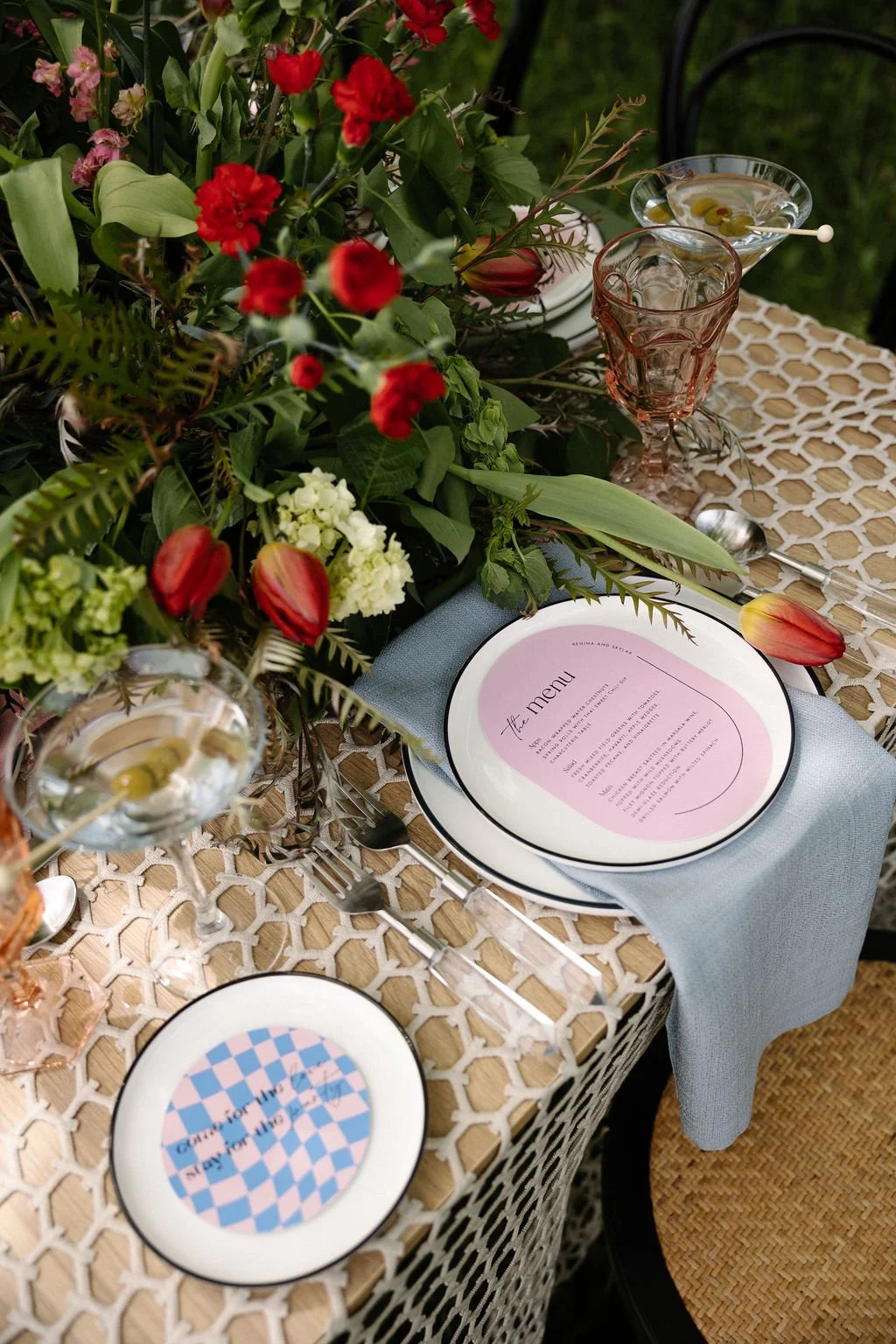 A decorated outdoor dining table with a large floral centerpiece consisting of red, pink, and white flowers with green foliage. The table is set with a light blue napkin, a pink and purple menu plate, a smaller plate with a message, and various glass