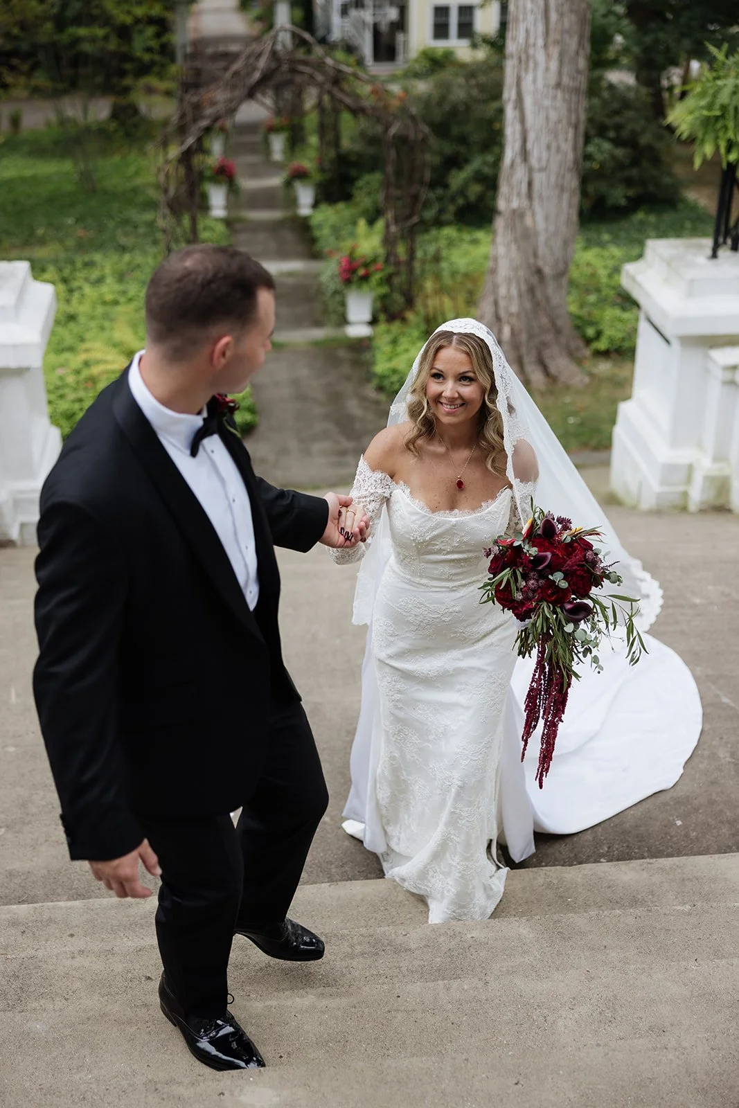 Bride and groom walking up stairs outdoors, holding hands. The bride is in a white wedding dress holding a bouquet of dark red flowers, and the groom is in a black tuxedo.