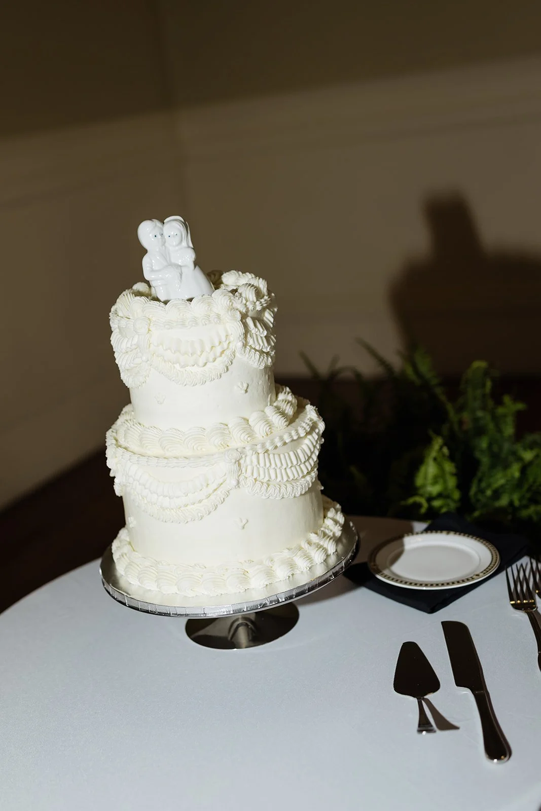 Three-tier white wedding cake with intricate piping decorations on a silver cake stand, topped with a wedding couple figurine. The cake is placed on a white table with a decorative plate, black napkin, and cake serving utensils beside it, in a settin