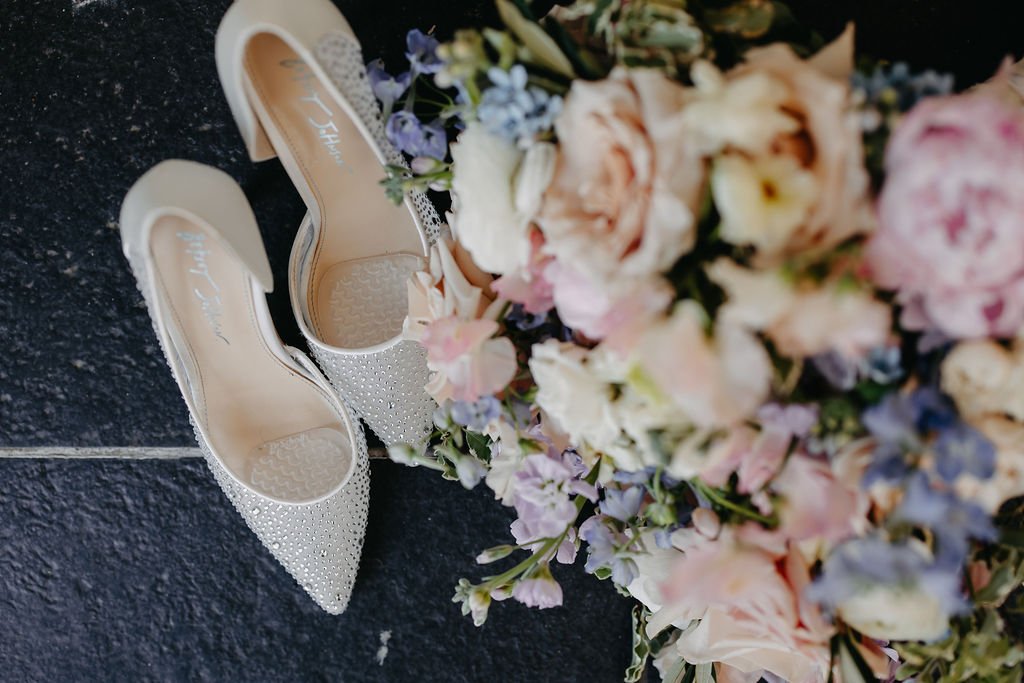 Pair of white high-heeled shoes with sparkling embellishments beside a large bouquet of pastel-colored flowers on a dark floor.