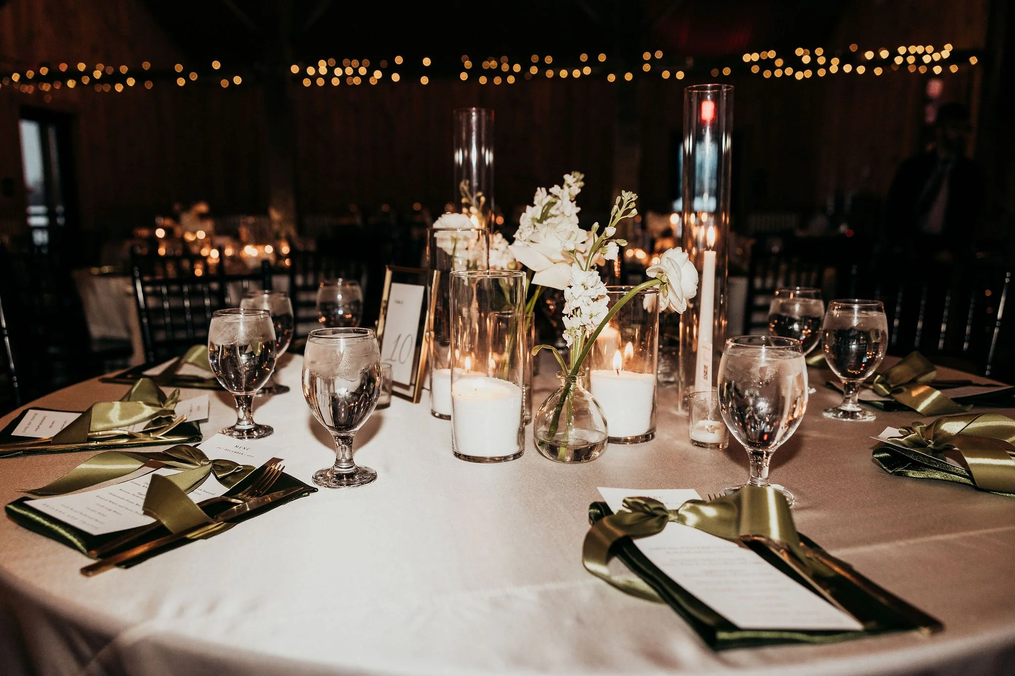 A decorated event table with white tablecloth, floral centerpieces, candles, and menus with gold ribbons in a dimly lit room with string lights.