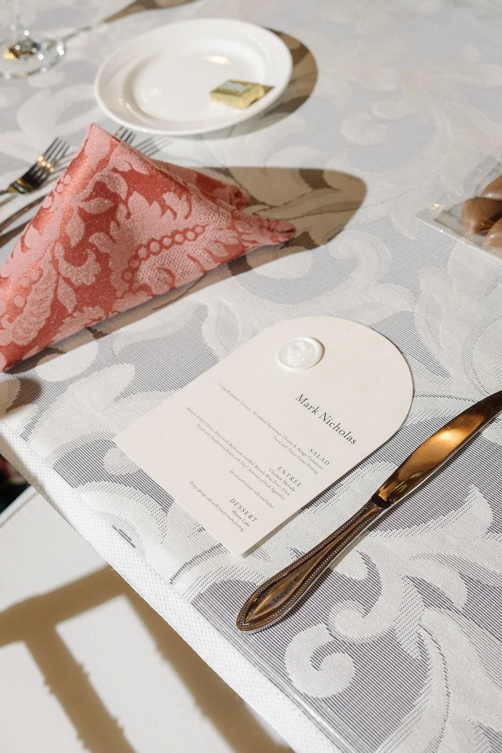 Dinner table setup with a menu, pink napkin, knife, and a plate with butter, decorated with a patterned tablecloth.