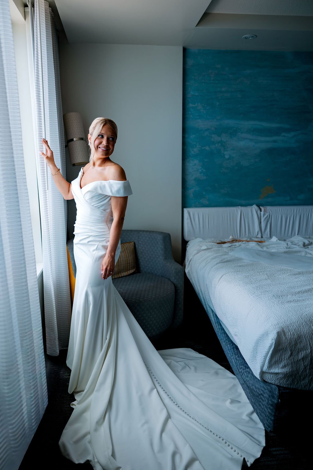 A woman in a white wedding dress standing by a window in a hotel room, smiling and looking outside. The room has a blue accent wall, a bed with white linens, and a blue armchair.