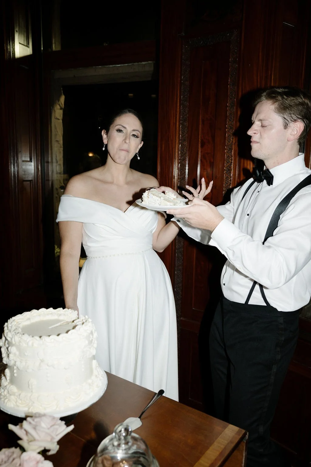 Bride in a white off-shoulder wedding dress with an expression of displeasure or disgust, holding a piece of wedding cake on a plate, being offered more cake by a man in a tuxedo, in a wooden-paneled room with a wedding cake on the table in front.