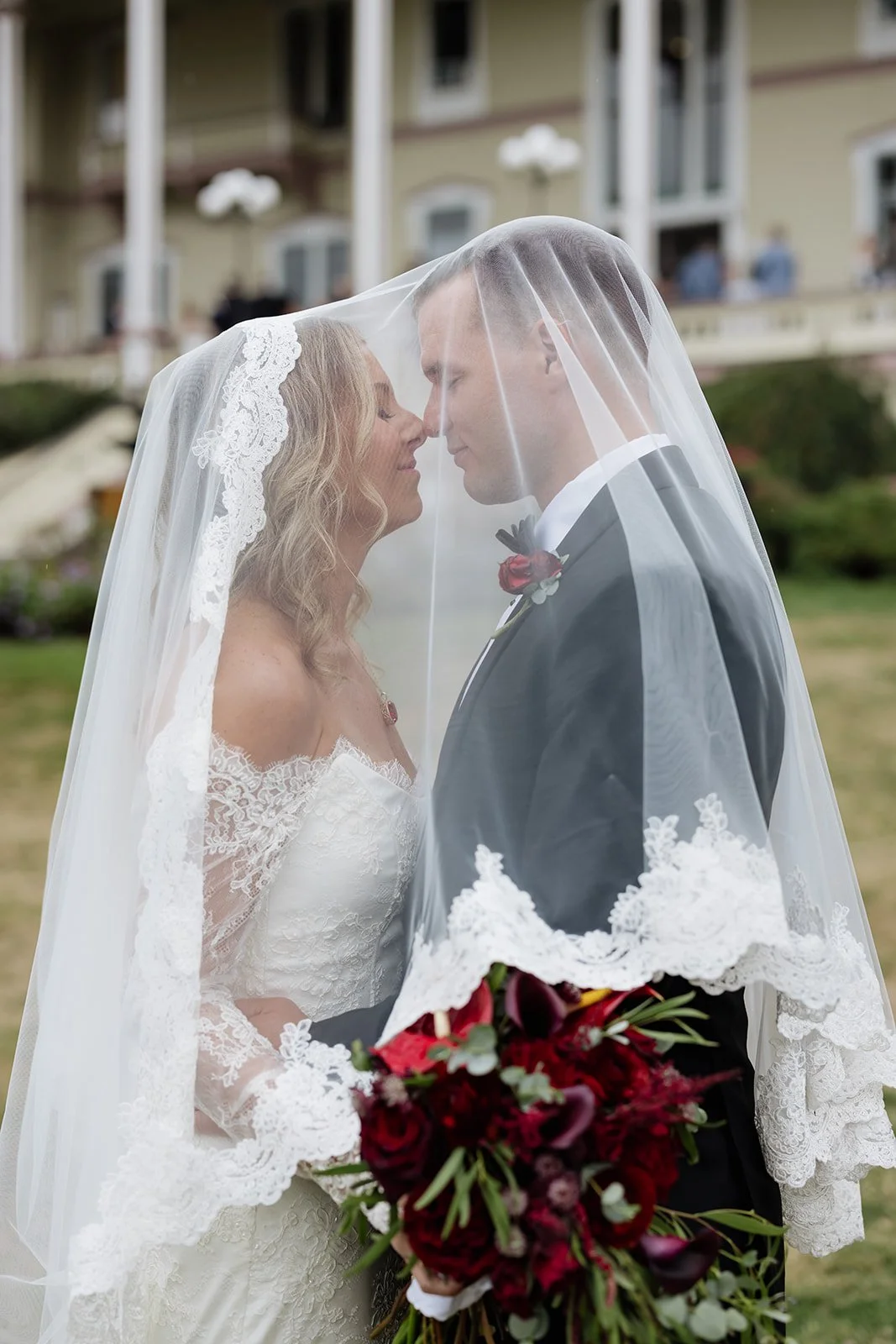 A bride and groom are touching foreheads, standing under a lace-edged veil, outside during a wedding. The bride is in a lace wedding dress, and the groom in a tuxedo with a red boutonniere, holding a bouquet of dark red flowers.