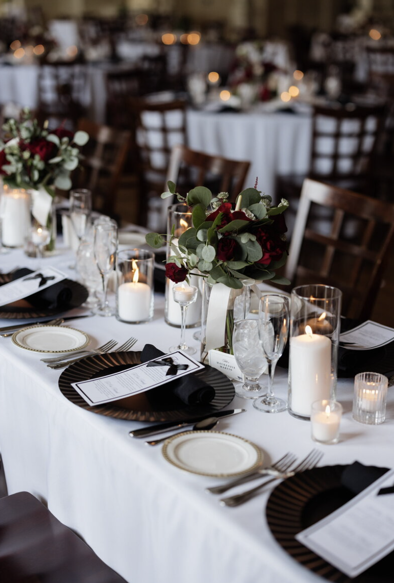 Elegant banquet table decorated with white tablecloth, black and gold plates, black napkins, and silverware. Centerpiece of red roses and greenery in a glass vase, surrounded by white and pillar candles in glass holders, with wine glasses and water glasses set for guests in a dimly lit venue.