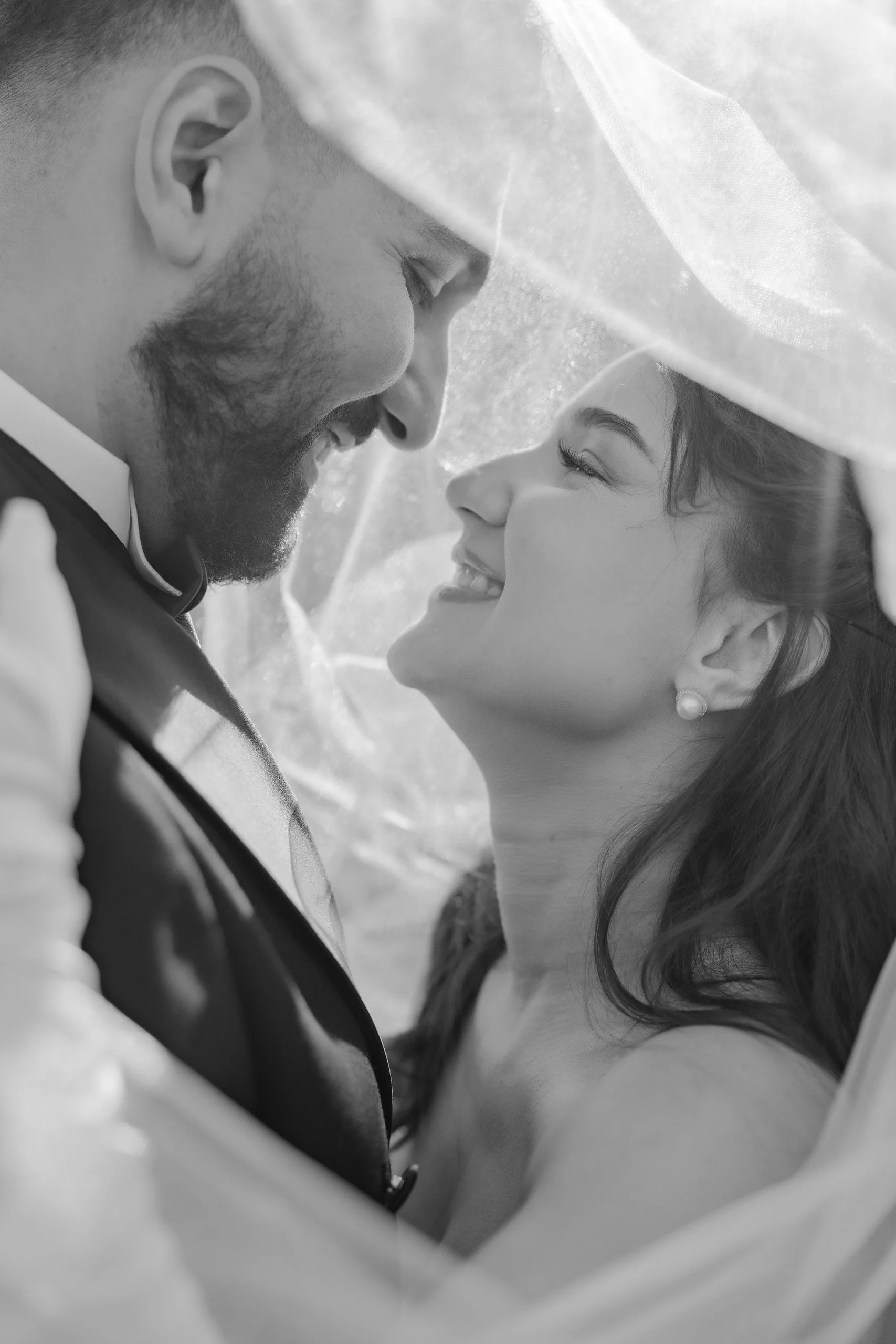 A black and white close-up photograph of a man and a woman smiling and touching foreheads, with the woman's face partially obscured by a veil or sheer fabric.
