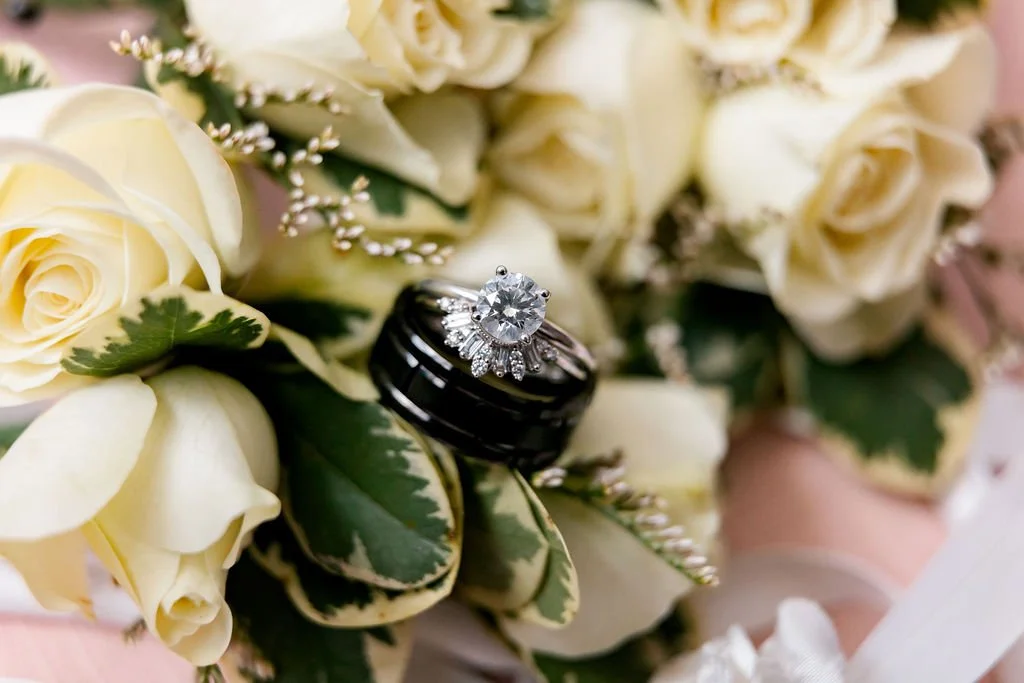 A close-up of a bouquet with white roses and greenery, featuring a diamond engagement ring and a black wedding band resting on the flowers.