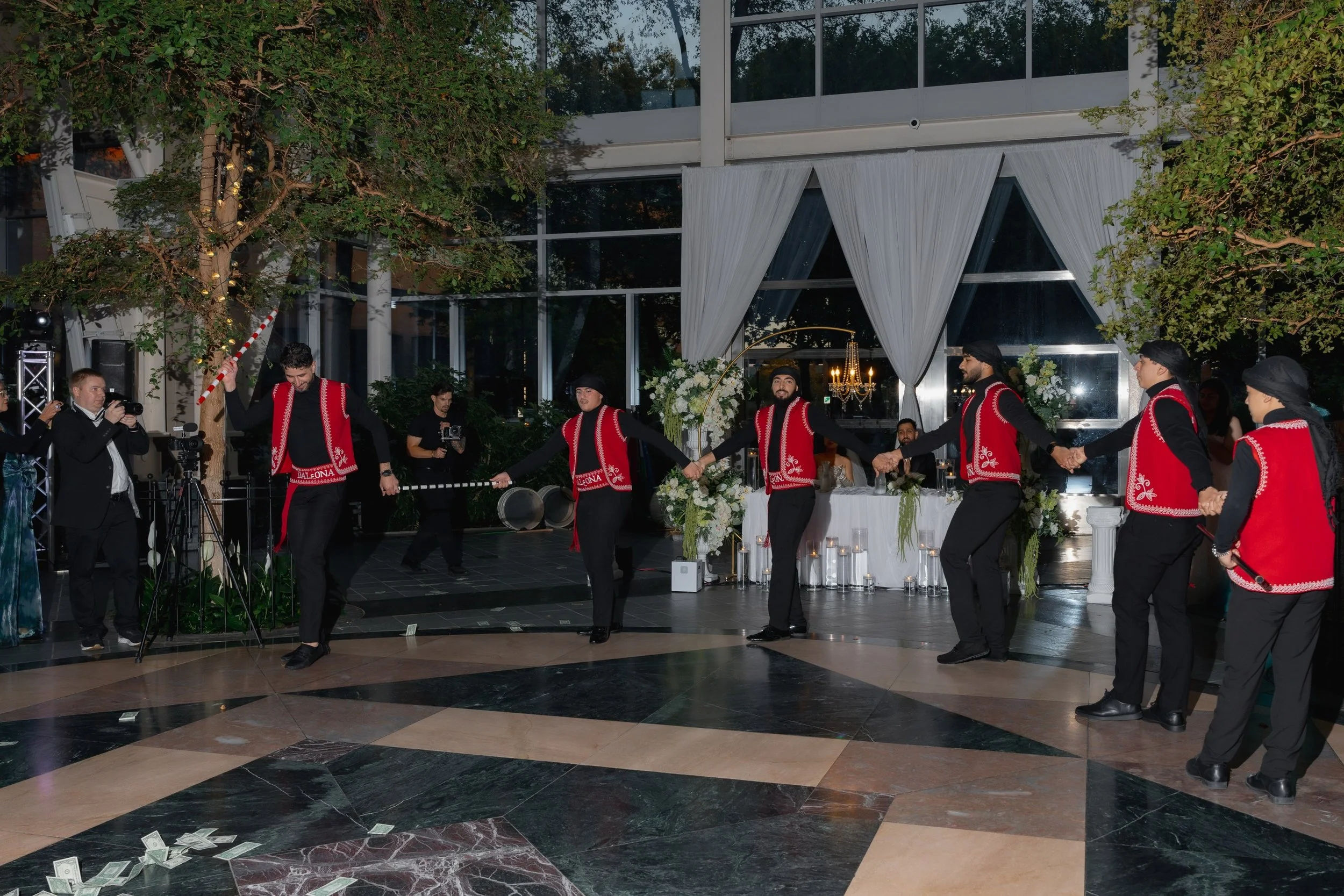 A group of men dressed in traditional black attire with red vests, holding hands and dancing in a circle at an indoor event with floral decorations and a chandelier.