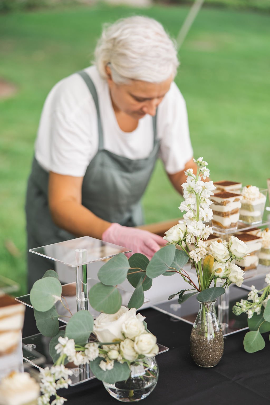 A woman with white hair arranging desserts on a table decorated with white flowers and green leaves in vases outdoors.