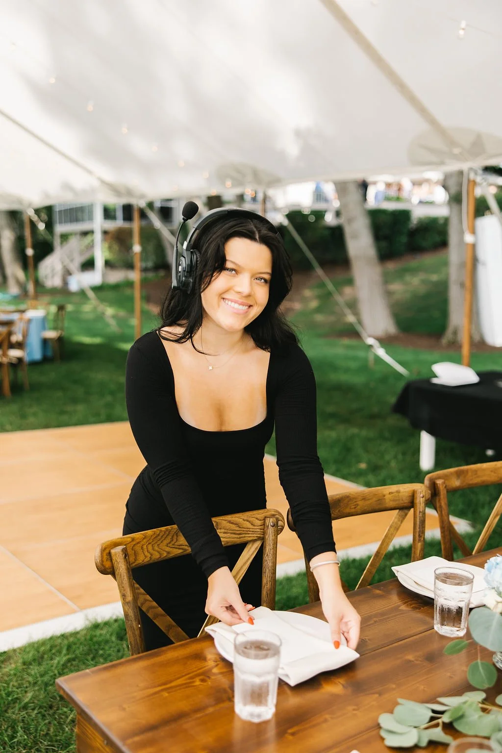 A smiling woman with long dark hair wearing a black top, standing inside a large white tent, preparing to set a dining table with folded napkin and glasses, on a sunny day at an outdoor event.