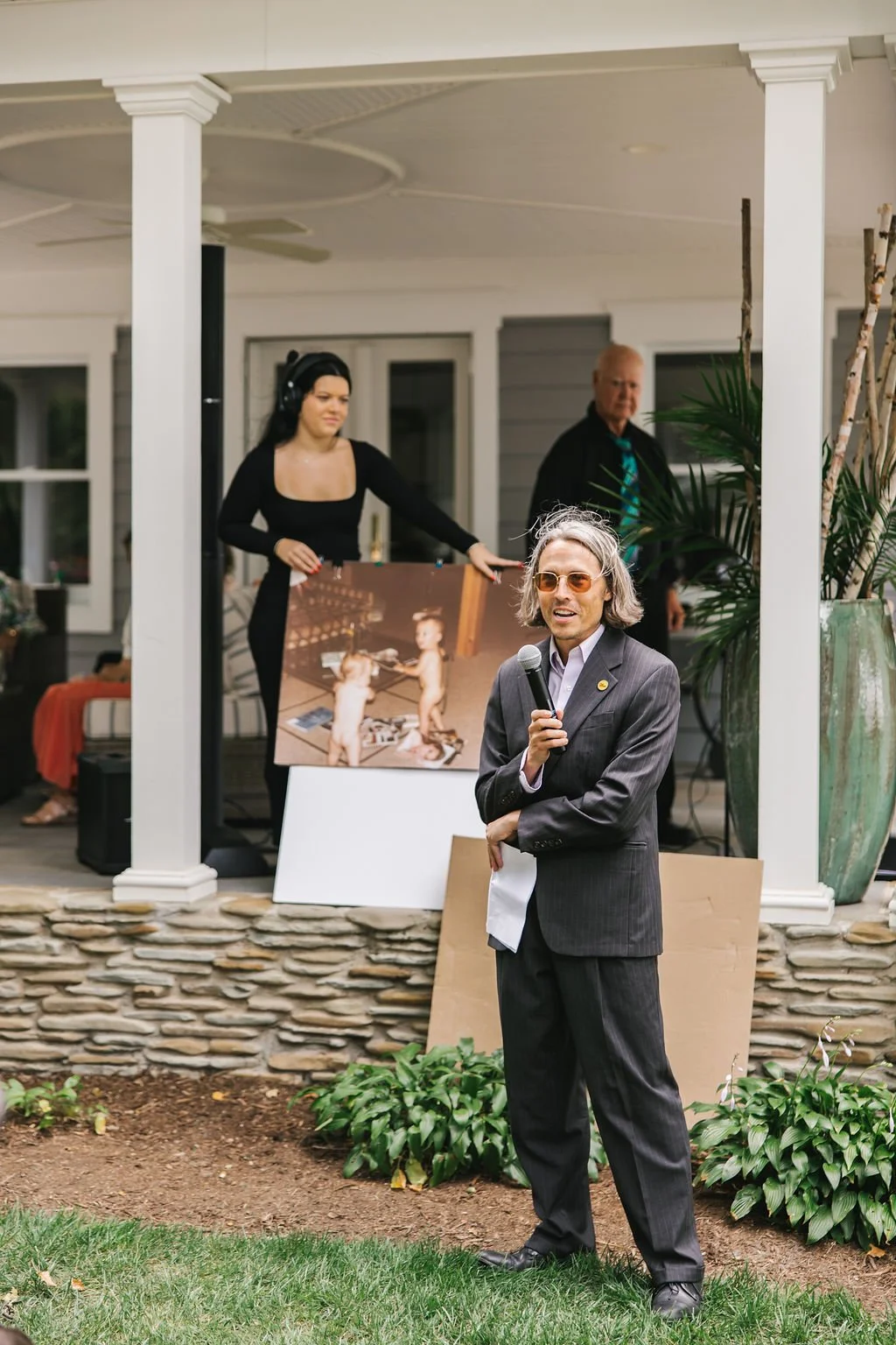 A man in a gray suit and sunglasses speaking into a microphone at an outdoor gathering, with a woman in black and a man in black behind him, and a large photo on an easel.