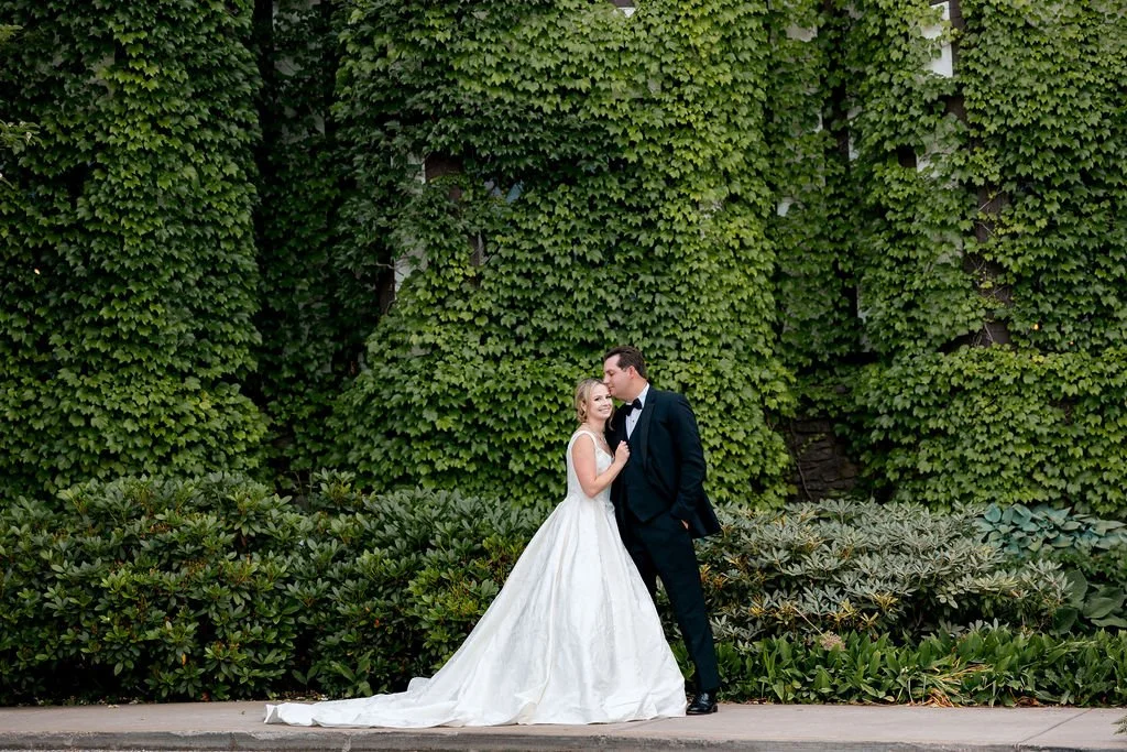 A bride and groom in wedding attire standing in front of a lush green ivy-covered wall.