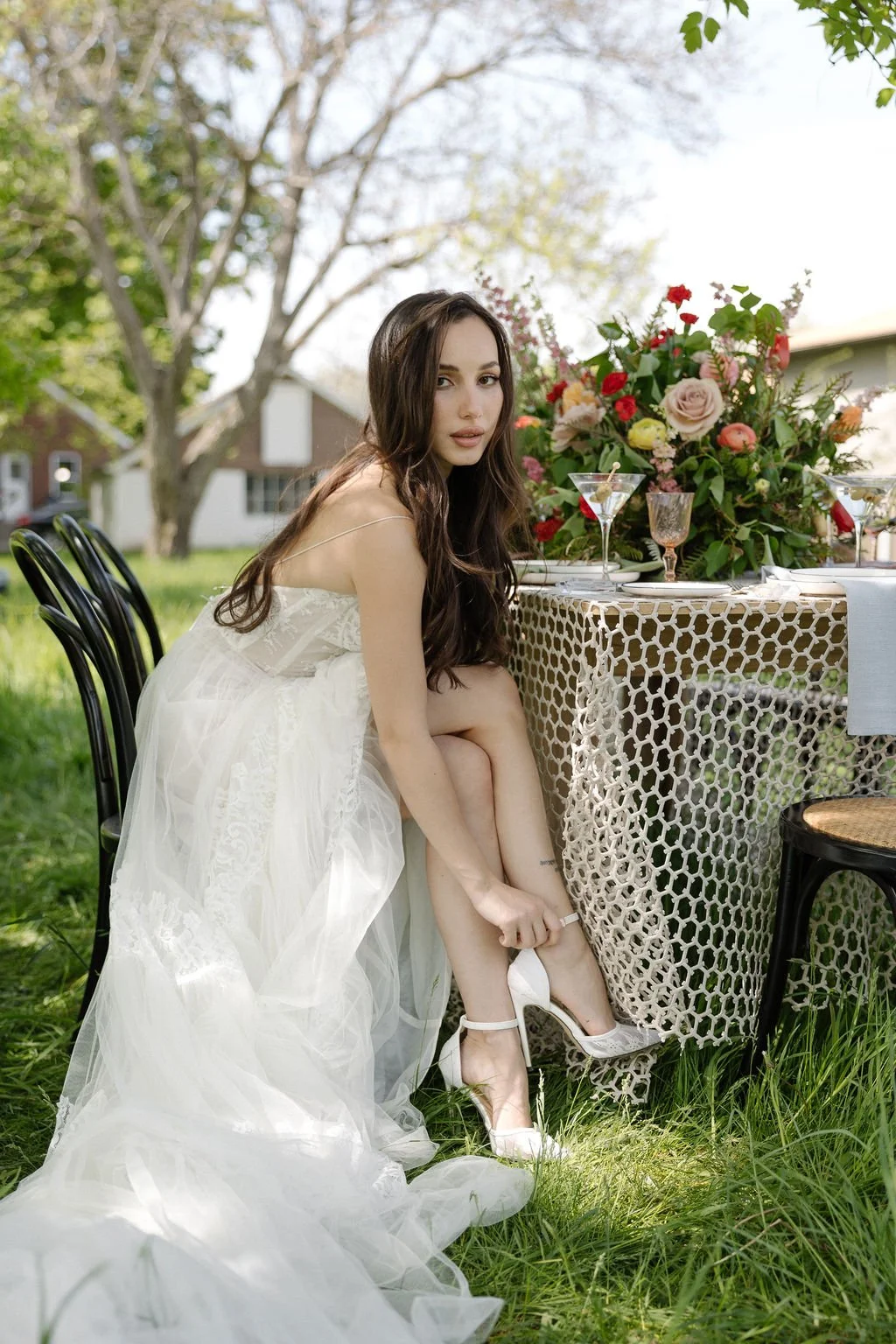 A woman in a white wedding dress sitting outside at a table with a floral centerpiece, martini glass, and other drinks, adjusting her white high-heeled shoe on her foot.