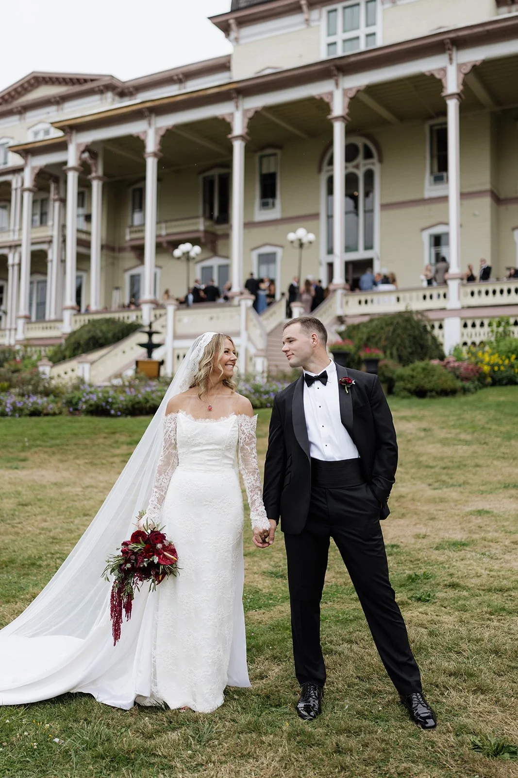 A bride and groom holding hands on a grassy lawn, smiling at each other. The bride is wearing a white lace wedding gown with a long veil, holding a bouquet of red flowers. The groom is in a black tuxedo with a bow tie. In the background, there is a l