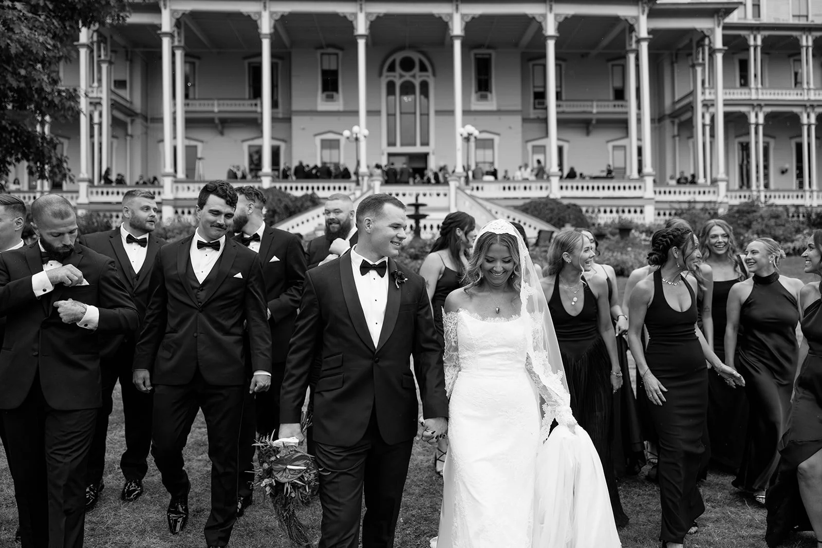 Black and white photo of a wedding celebration with a bride and groom walking hand in hand and surrounded by wedding guests in formal attire, in front of a large, ornate building.
