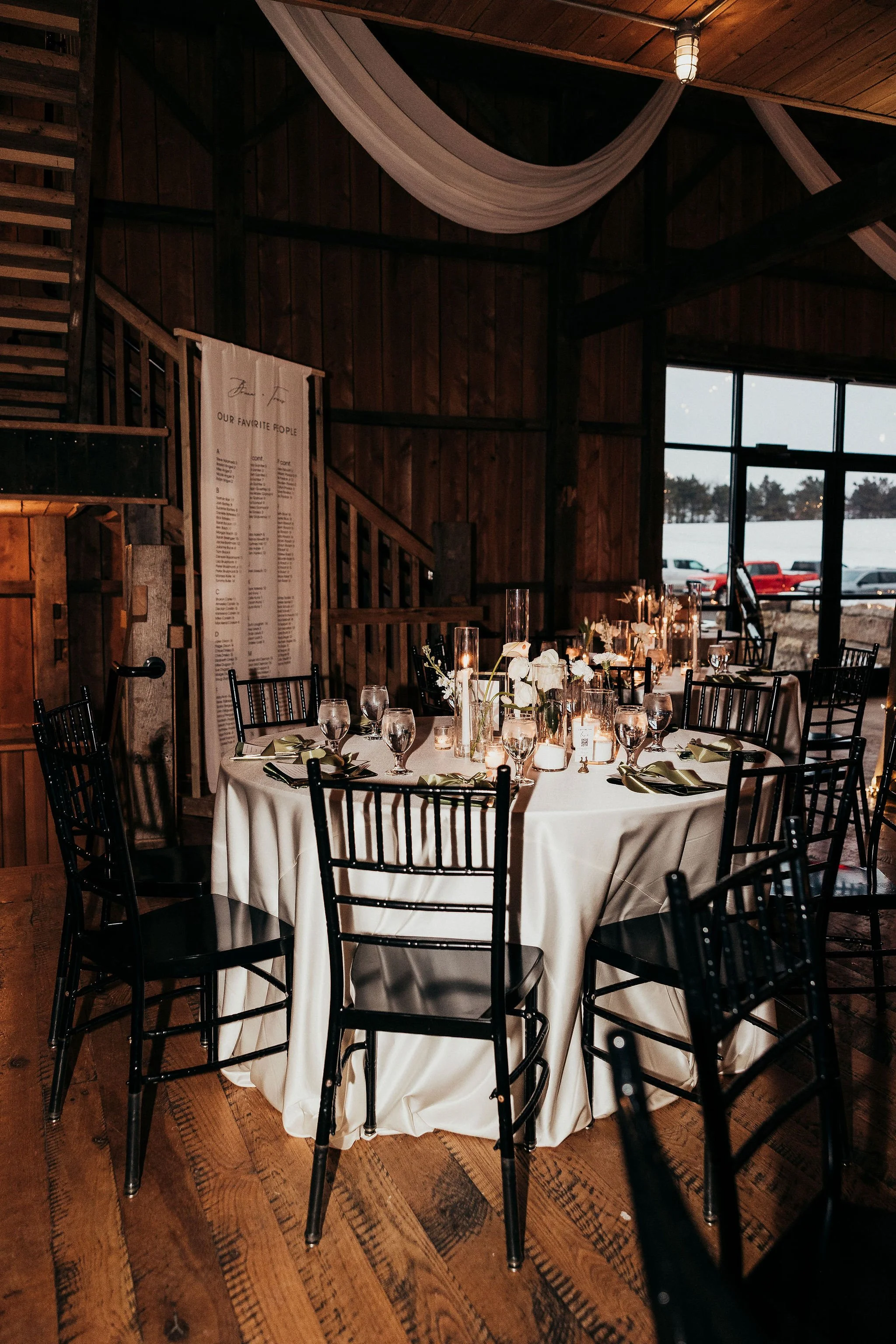 Indoor banquet or event hall with a round table set for a formal meal. The table has a white tablecloth, candles, and floral centerpieces. Wooden walls and ceiling, with large windows showing a snowy landscape and parked cars outside.