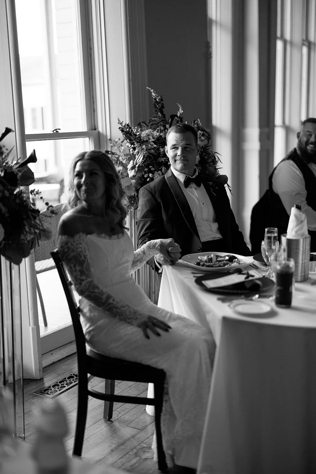 Black and white photo of a bride and groom seated at a table during a wedding reception. The bride is wearing a lace wedding dress and holding the groom’s hand. The groom is in a tuxedo, holding a plate of food. Other guests and floral decorations ar