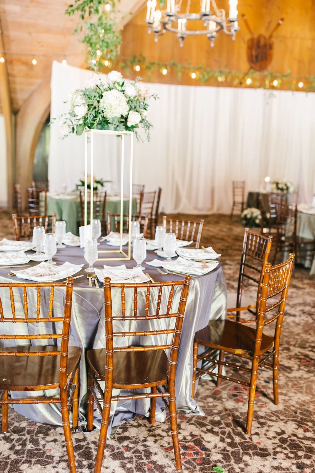 Round banquet table set for a formal event with a tall floral centerpiece, white napkins, and glassware, in a rustic event space with wood walls, string lights, and additional decorated tables in the background.