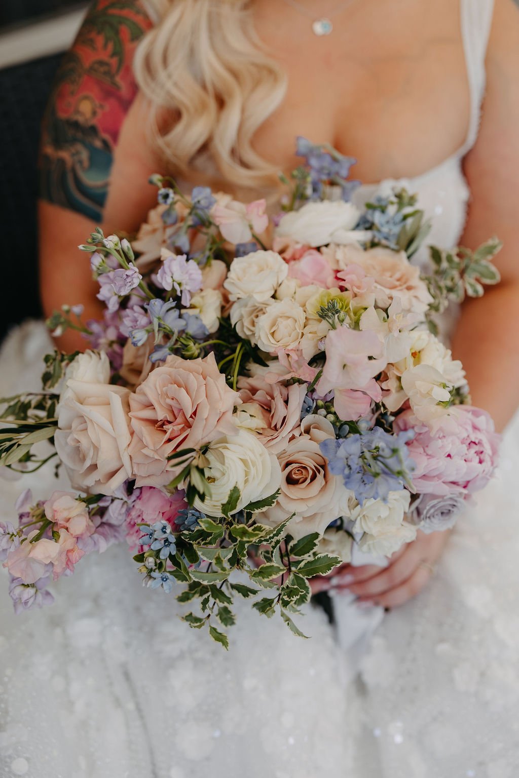 A woman in a wedding dress holding a large bouquet of pastel-colored flowers, including roses and delphiniums.