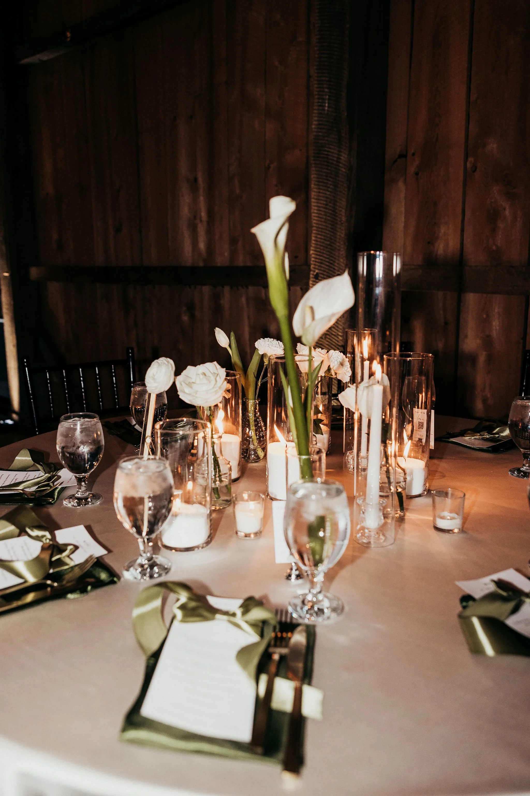A dining table set for a formal event with candles, white flowers, glasses of water, and gold ribbon napkin holders on a beige tablecloth, against a dark wooden wall.