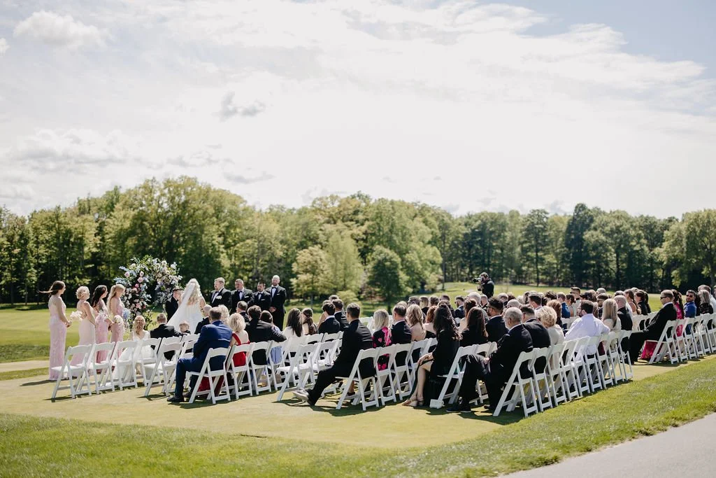 Outdoor wedding ceremony with seated guests, bride and groom at the altar, officiant, and bridesmaids in pink dresses, set in a green landscape with trees and a cloudy sky.