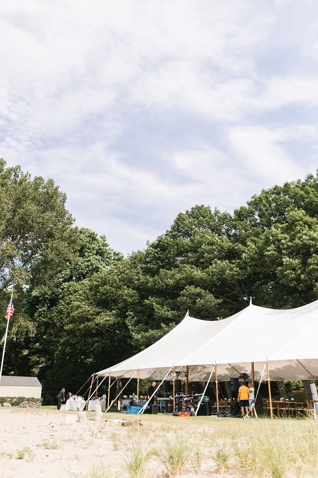 A white event tent set up outdoors on a grassy area with multiple people preparing equipment underneath. There are large trees in the background and an American flag on a pole to the left.